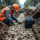 Bouwvakker in oranje vest onderzoekt bioswale en plastic kratmodules in sleuf, meetlint in hand op Nederlandse bouwplaats