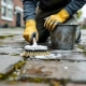 Persoon schrobt grijze betonnen tegels schoon met borstel en zeepsop, draagt werkkleding en handschoenen op Nederlandse straat