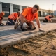 Arbeiders leggen grijze betonklinkers op industrieterrein, rubber hamer in actie, zandbed zichtbaar, oranje veiligheidsvesten, gouden uurtlicht.