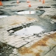 Beschadigde parkeerplaats met gebarsten bestrating, ongelijke asfaltpatches en plassen regenwater, oranje verkeerskegels markeren gevaarlijke zones.