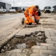 Medewerker in oranje veiligheidsvest inspecteert gebarsten en verzakte betonklinkers op een versleten industrieterreinweg.