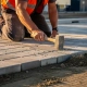 Vakman in oranje veiligheidsvest legt betonklinkers op zandbed in parkeerplaats, rubber hamer in hand, gouden middaglicht.