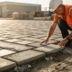 Bouwvakker in oranje veiligheidsvest inspecteert verse betonklinkers op Nederlands industrieterrein, geknield in middagzon.