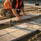 Vakman in oranje veiligheidsvest legt betonnen straatklinkers op zandbed bij bedrijventerrein, rubberen hamer op de grond, gouden uurtje.