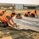 Arbeiders in oranje vesten leggen grijze betonklinkers op een zandbedding in een industrieel parkeerterrein tijdens gouden avondlicht.