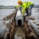 Arbeiders in gele veiligheidsvesten leggen een betonnen afwateringskanaal aan in een parkeerplaats, met modderige greppel en grijze bestrating.