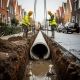 Arbeiders in gele hesjes plaatsen betonnen afvoerbuis in modderige bouwput naast nieuw bestraatwerk in Nederlandse woonstraat.