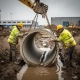 Twee bouwvakkers in gele hesjes begeleiden een betonnen drainagepijp die door een graafmachine in een modderige bouwput wordt neergelaten.