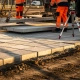 Arbeiders plaatsen betonnen straatplaten op bedrijventerrein met hijsapparatuur, meetapparatuur en GPS zichtbaar in middagzon.