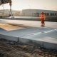 Betonplaten worden precisie gelegd op een industrieterrein, begeleid door een arbeider in oranje veiligheidsvest, met hijsmachine en zandondergrond op de achtergrond.