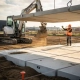 Betonnen parkeerplaatsplaten geplaatst door minigraver op Nederlands bouwterrein, arbeider in oranje veiligheidsvest stuurt plaatsing aan.