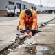 Veiligheidsmedewerker in oranje vest inspecteert gescheurde betonvloer op Nederlands industrieterrein, geknield bij voegerosie tussen betonplaten.