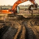 Oranje graafmachine graaft kleigrond op een bedrijventerrein in ontwikkeling, met uitzetpalen en lange schaduwen in de late middagzon.