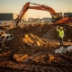 Oranje graafmachine breekt kleigrond op Nederlands bouwterrein, veiligheidsmedewerker in geel vest begeleidt werkzaamheden, betonnen drainagebuizen op de achtergrond.