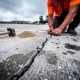 Inspecteur in oranje veiligheidsvest onderzoekt gebarsten voegwerk op verweerd betonnen parkeerterrein, rubber hamer en voegzand nabij.