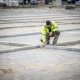 Medewerker in geel veiligheidsvest inspecteert betonstraatstenen van een nieuw aangelegde parkeerplaats onder bewolkt Nederlands daglicht.