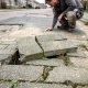 Vakman inspecteert verzakte en gebarsten stoeptegels met onkruid in voegen op een Nederlandse woonstraat.
