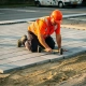 Stratenmaker in oranje veiligheidsvest legt grijze betonklinkers op zandbed in commerciële parkeerplaats, rubber hamer in hand.
